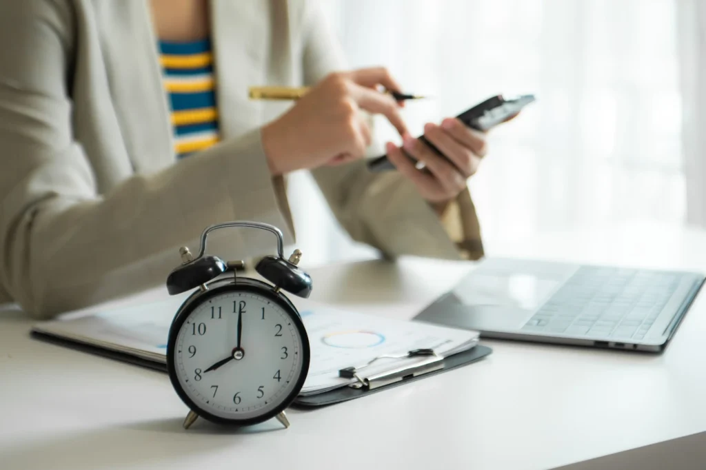 Person using smartphone with alarm clock, notebook, and laptop on desk.